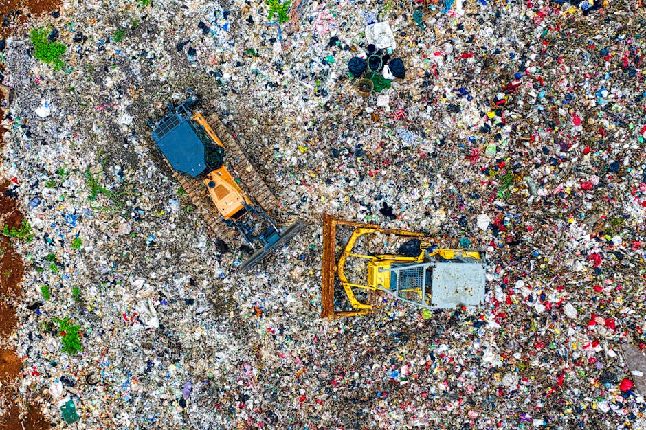 An aerial view of a large landfill site filled with scattered mixed waste, including plastic bottles, paper, textiles, and household items. Two heavy-duty excavators with hydraulic arms and buckets are actively engaged in managing the debris; one is orange with tracks, and the other is yellow and grey with a metal bucket, both positioned within the refuse pile. The surrounding area consists of compacted waste and loose materials, with some patches of green vegetation visible at the edges. The scene captures the scale of waste disposal operations, which are part of the broader waste management processes supporting house removals and disposal services, as provided by Man With a Van Primrose Hill during relocation and debris clearance activities.