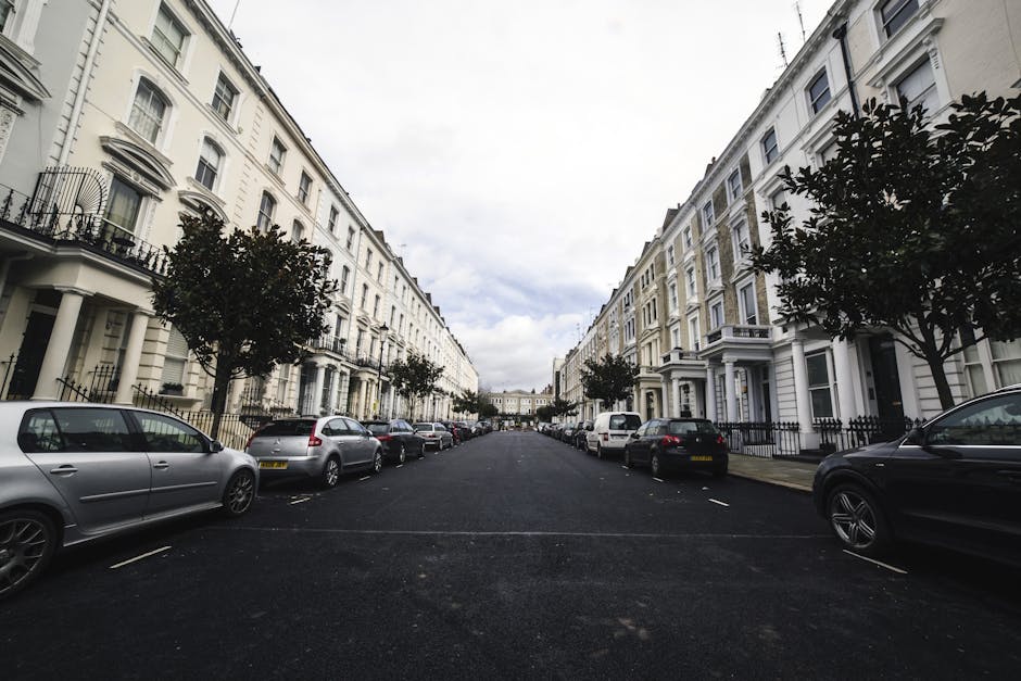 A view of a residential street with white terraced Victorian-style houses on both sides, each featuring black wrought iron railings and bay windows. Several trees line the pavement, providing some greenery amid the closely arranged buildings. Parked cars, including compact and hatchback models in silver, black, and grey, are positioned along the curb in front of the residences. The street surface is paved with dark asphalt, and the sky above is partly cloudy with patches of light blue. This scene depicts a typical urban environment suitable for house removals, with the quiet street illustrating the logistics involved in home relocation and furniture transport. Occasionally, moving services such as those offered by Man With a Van Primrose Hill might utilize this street for loading and unloading furniture during packing and moving operations, often involving vehicle access and street permits, especially for larger items or multiple trips.