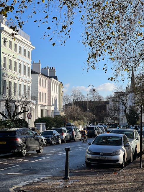 A view of a residential street with white terraced Victorian-style houses on both sides, each featuring black wrought iron railings and bay windows. Several trees line the pavement, providing some greenery amid the closely arranged buildings. Parked cars, including compact and hatchback models in silver, black, and grey, are positioned along the curb in front of the residences. The street surface is paved with dark asphalt, and the sky above is partly cloudy with patches of light blue. This scene depicts a typical urban environment suitable for house removals, with the quiet street illustrating the logistics involved in home relocation and furniture transport. Occasionally, moving services such as those offered by Man With a Van Primrose Hill might utilize this street for loading and unloading furniture during packing and moving operations, often involving vehicle access and street permits, especially for larger items or multiple trips.