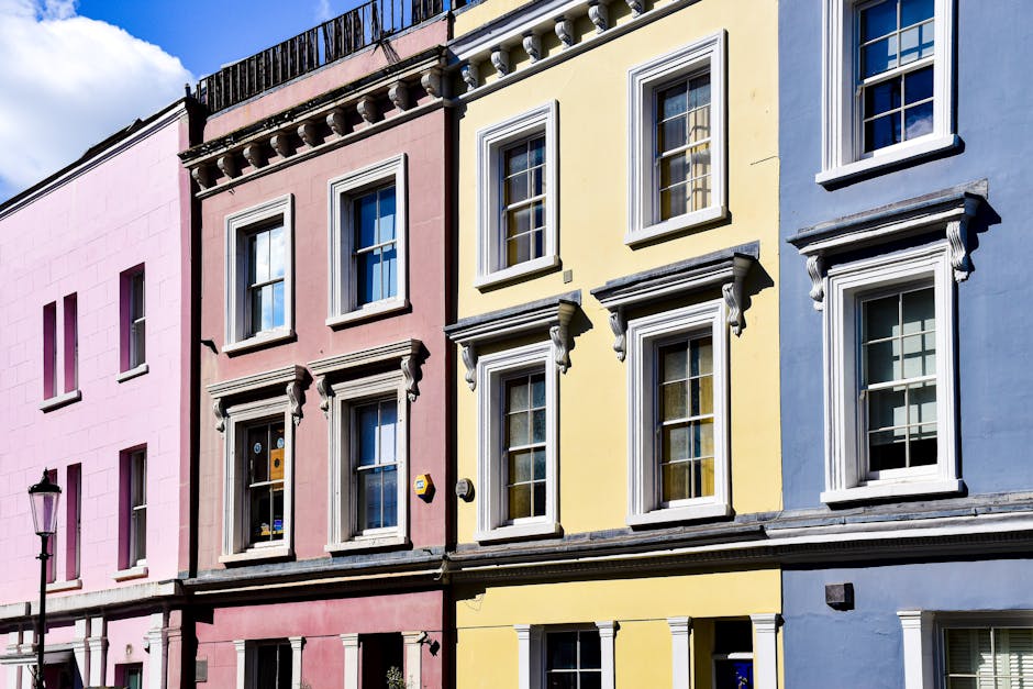 A row of three pastel-coloured terraced houses with large white-framed sash windows and decorative cornices, situated on a street in Primrose Hill. The pink, yellow, and blue facades each feature multiple floors with evenly spaced windows, some open revealing interior curtains. The buildings are illuminated by bright daylight, with a clear blue sky overhead. On the left, a traditional black street lamp stands on the pavement. The image is related to house removals and moving services offered by Man With a Van Primrose Hill, highlighting the typical residential environment involved in home relocation and furniture transport processes.