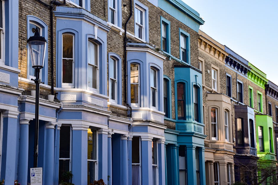 A row of three pastel-coloured terraced houses with large white-framed sash windows and decorative cornices, situated on a street in Primrose Hill. The pink, yellow, and blue facades each feature multiple floors with evenly spaced windows, some open revealing interior curtains. The buildings are illuminated by bright daylight, with a clear blue sky overhead. On the left, a traditional black street lamp stands on the pavement. The image is related to house removals and moving services offered by Man With a Van Primrose Hill, highlighting the typical residential environment involved in home relocation and furniture transport processes.