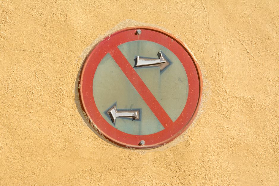 A round traffic sign mounted on a yellow wall, indicating no left or right turn with a red circle and diagonal line. The sign has a greyish-green background and features two protruding metal arrows pointing left and right, both of which are bent and attached to the sign. The sign appears weathered with some peeling paint around the edges. The setting suggests an exterior environment near a property or street, possibly adjacent to a loading area or pavement used for house removals. This type of signage is relevant in the context of logistics and moving operations, such as those provided by Man With a Van Primrose Hill, especially when adhering to local loading bay rules, as indicated by the page title.