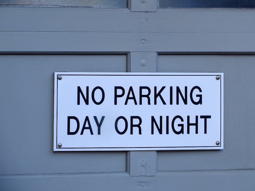 A rectangular white sign with bold black text reading 'NO PARKING DAY OR NIGHT' is mounted on a light grey wall, likely outside a residential or commercial property in NW1, Primrose Hill. The sign is secured with four metallic screws at each corner. The wall above the sign features horizontal wooden panels, painted in a matching grey color. This setting appears to be in an urban environment, possibly near a loading bay or vehicle access point used by house removals or furniture transport services. The image emphasizes the importance of observing parking regulations during home relocation or moving activities, as managed by Man With a Van Primrose Hill, which provides professional removals and packing services, especially when planning deliveries or loading during scheduled moving logistics.