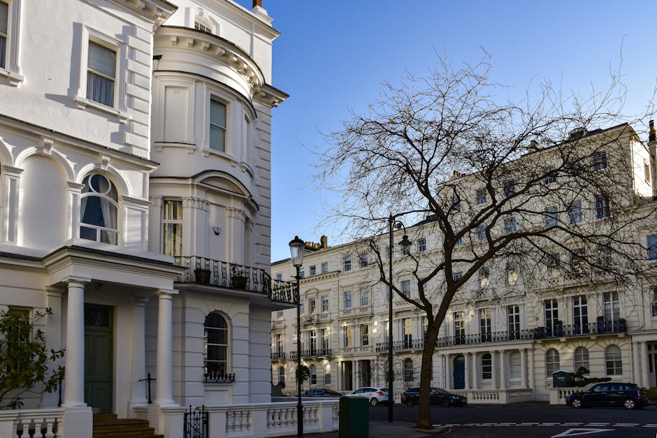 A street scene in Primrose Hill with classic Georgian-style terraced houses lining the left side, featuring tall windows, decorative cornices, and wrought iron balconies. Several medium-sized cardboard boxes and wrapped furniture pieces are positioned on the pavement near the building entrances, indicating packing for home relocation. A person is seen lifting a box onto a trolley close to the doorway, while another stands nearby, possibly giving instructions. A black van with its sliding door open is parked on the road, ready for furniture transport, with protective blankets draped over some items inside. Additional cars are parked along the street, which is shaded by mature trees, and the sky above is clear with a few clouds. This scene illustrates a house removal operation in progress within the Primrose Hill area, involving careful packing, loading, and vehicle preparation by professionals such as Man With a Van Primrose Hill for efficient moving services. The environment reflects a typical urban residential setting suitable for appropriate logistics and moving procedures.