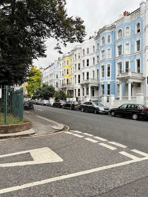 The image shows a street scene in Primrose Hill with a row of white and pastel blue Victorian-style residential buildings featuring bay windows and decorative facades. Parked cars line the side of the quiet road, which has a designated parking bay and a pedestrian crossing with white dashed lines. On the left, a large leafy tree partly obscures a green metal fence surrounding a small garden area. The pavement is made of grey slabs, and there is a curb separating the sidewalk from the roadway. Overcast skies cast diffuse lighting over the scene. This setting reflects an urban residential area suitable for house removals or home relocation services, with a focus on urban furniture transport, loading, and parking logistics, which are common elements in the context of professional removals like those provided by Man With a Van Primrose Hill.