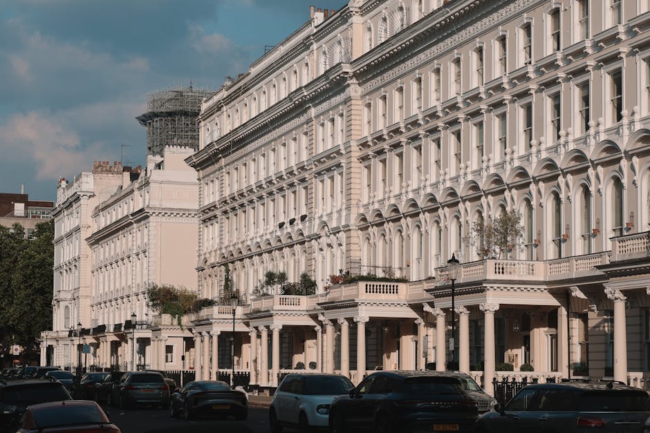 A row of elegant white terraced townhouses along a busy urban street with parked cars lining the curb. The buildings feature large windows with decorative molding, small balconies with balustrades, and some have potted plants on the terraces. In the foreground, several cardboard boxes, wrapped furniture, and moving blankets are visible on the pavement near the entrance of one property, indicating a house relocation process. A man with a van is loading or unloading items onto a vehicle parked on the street, with visible moving straps and trolleys nearby. The scene is well-lit under partly cloudy skies, with the presence of a van from Man With a Van Primrose Hill subtly supporting local removals and transportation services related to packing and moving. The environment suggests an active home relocation or furniture transport operation in an urban setting, with attention to careful handling and loading of household items into the van.