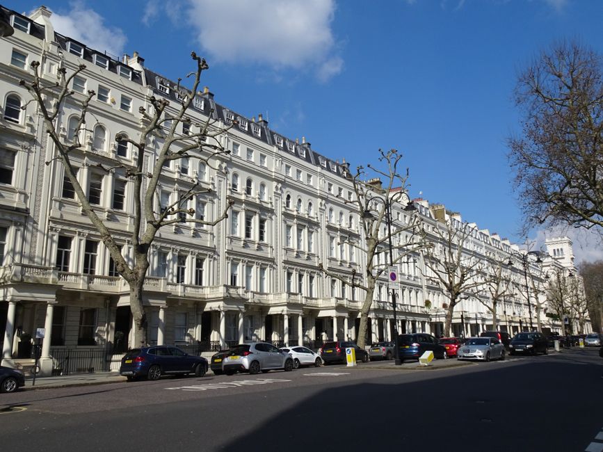 The image shows a street scene in Primrose Hill with a row of white and pastel blue Victorian-style residential buildings featuring bay windows and decorative facades. Parked cars line the side of the quiet road, which has a designated parking bay and a pedestrian crossing with white dashed lines. On the left, a large leafy tree partly obscures a green metal fence surrounding a small garden area. The pavement is made of grey slabs, and there is a curb separating the sidewalk from the roadway. Overcast skies cast diffuse lighting over the scene. This setting reflects an urban residential area suitable for house removals or home relocation services, with a focus on urban furniture transport, loading, and parking logistics, which are common elements in the context of professional removals like those provided by Man With a Van Primrose Hill.
