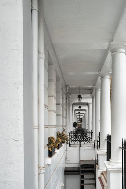 A long, narrow exterior corridor with white painted stone walls and pillars supporting an overhead balcony. The corridor features decorative black iron railings, planters with dried plants, and black lantern-style wall-mounted lights. At the far end, a dark doorway is visible. The setting appears to be part of a residential building in Primrose Hill, with the corridor facilitating access and movement. It is daytime, with natural light illuminating the space, which may be used for a home relocation or furniture transport process, as indicated by context related to house removals. The architectural style and compact layout suggest challenges in moving large items, such as furniture, in narrow spaces typical of Primrose Hill properties, aligning with the theme of staircase and narrow lane challenges in house moves. Man With a Van Primrose Hill, a professional removals company, might service such environments during packing and loading procedures.