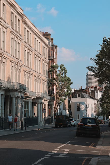 A daytime street scene in Primrose Hill featuring a row of multi-storey residential buildings with light-colored facades, decorative window frames, and balconies. Several trees line the sidewalk, some with visible foliage and branches extending over parked cars. On the pavement, a few pedestrians are walking, and there are parked vehicles including a black van, which is partially visible, suggesting a residential or commercial area. The sky is clear with a few clouds, and the overall scene represents an urban environment suitable for house removals and local moving services, such as those provided by Man With a Van Primrose Hill. The image captures a typical street where home relocation or furniture transport might take place, with the evening light casting gentle shadows.